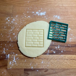 Puede incluir: Un cortador de galletas de plástico verde con forma de pared de ladrillo, sobre una masa de galletas redonda.