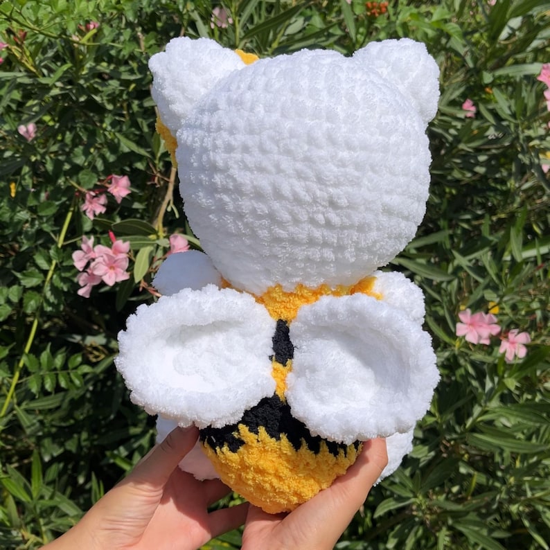 Puede incluir: Juguete de peluche de abeja blanca de crochet con rayas amarillas y negras y alas blancas. La abeja est&aacute; siendo sostenida en la mano de una persona.