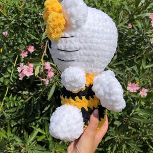 Puede incluir: Un juguete de peluche blanco tejido a crochet con forma de abeja con un cuerpo a rayas amarillo y negro. El juguete tiene una cara blanca y esponjosa con ojos negros y un lazo amarillo en la parte superior de la cabeza.