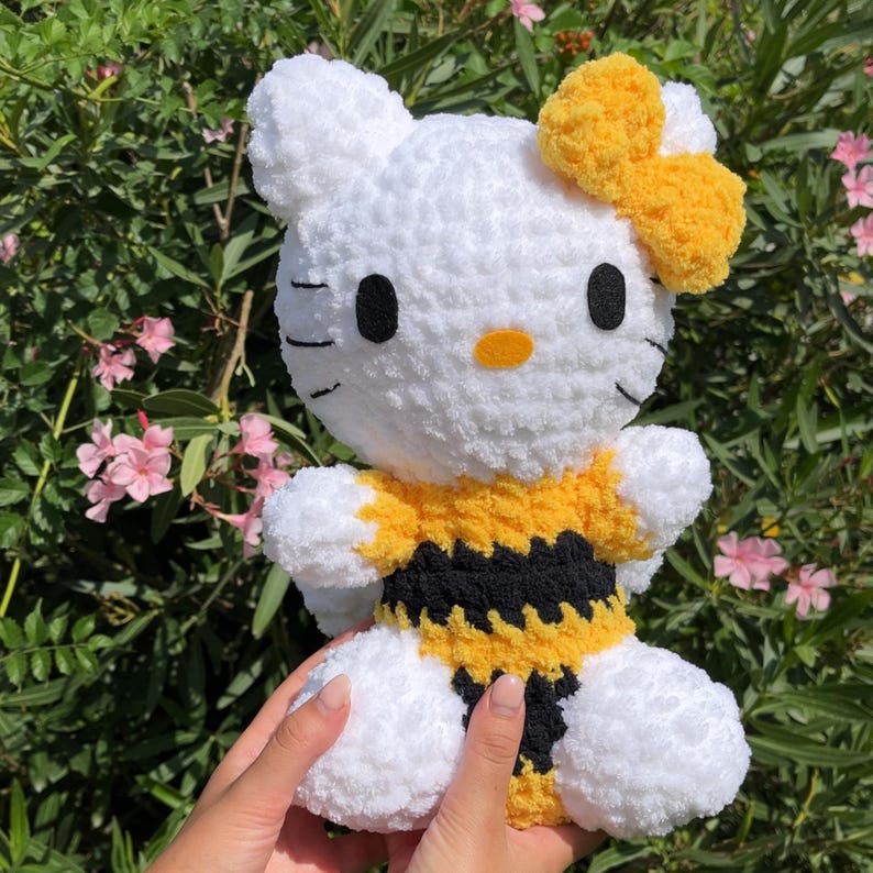 Puede incluir: Peluche Hello Kitty de crochet blanco con un disfraz de abeja amarillo y negro con un lazo amarillo en la cabeza.