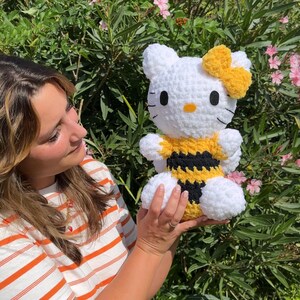 Puede incluir: Un juguete de peluche Hello Kitty blanco tejido a crochet con un disfraz de abeja amarillo y negro. El juguete tiene un lazo amarillo en la cabeza y ojos negros. El juguete est&aacute; siendo sostenido por una persona.