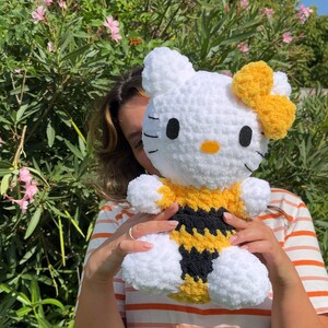 Puede incluir: Un juguete de peluche Hello Kitty blanco tejido a crochet con un disfraz de abeja amarillo y negro. El juguete tiene un lazo amarillo en la cabeza y ojos negros.