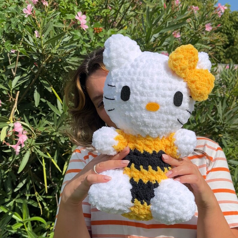 Puede incluir: Un juguete de peluche Hello Kitty blanco tejido a crochet con un disfraz de abeja amarillo y negro. El juguete tiene un lazo amarillo en la cabeza y ojos negros.