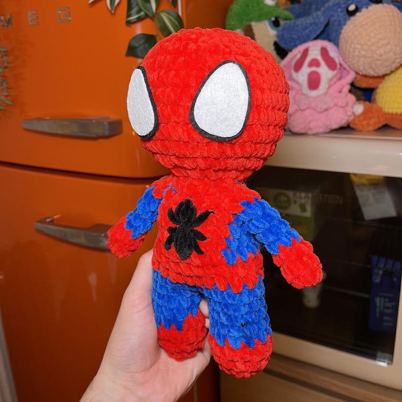 May include: A crocheted plush toy of Spider-Man in red and blue with a black spider symbol on the chest. The toy has white eyes and is holding its arms out.