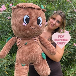 Pode incluir: Um brinquedo de pelúcia de crochê marrom que se assemelha ao personagem Groot da franquia de filmes Guardiões da Galáxia. O brinquedo tem uma cabeça grande com um rosto amigável, olhos grandes e um pequeno sorriso. O brinquedo está segurando uma folha verde na mão. O texto "PDF PATTERN ONLY" é visível em um coração rosa.