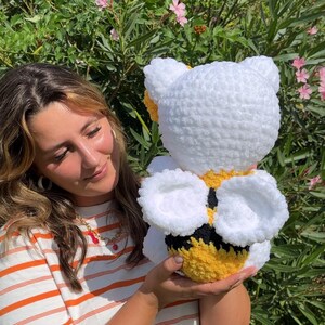 Puede incluir: Un juguete de peluche de abeja blanco tejido a crochet con rayas amarillas y negras y alas blancas. La abeja est&aacute; siendo sostenida por una persona que lleva una camisa a rayas.