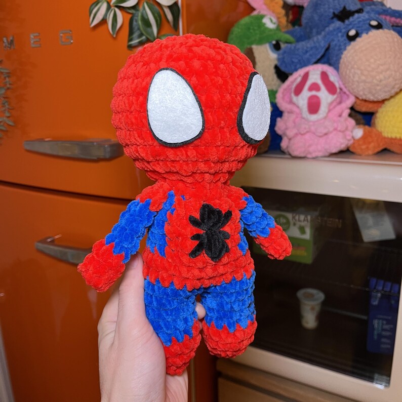 May include: A crocheted plush toy of Spider-Man. The toy is red and blue with a black spider symbol on the chest. The toy has white eyes and is holding a pose with its arms outstretched.