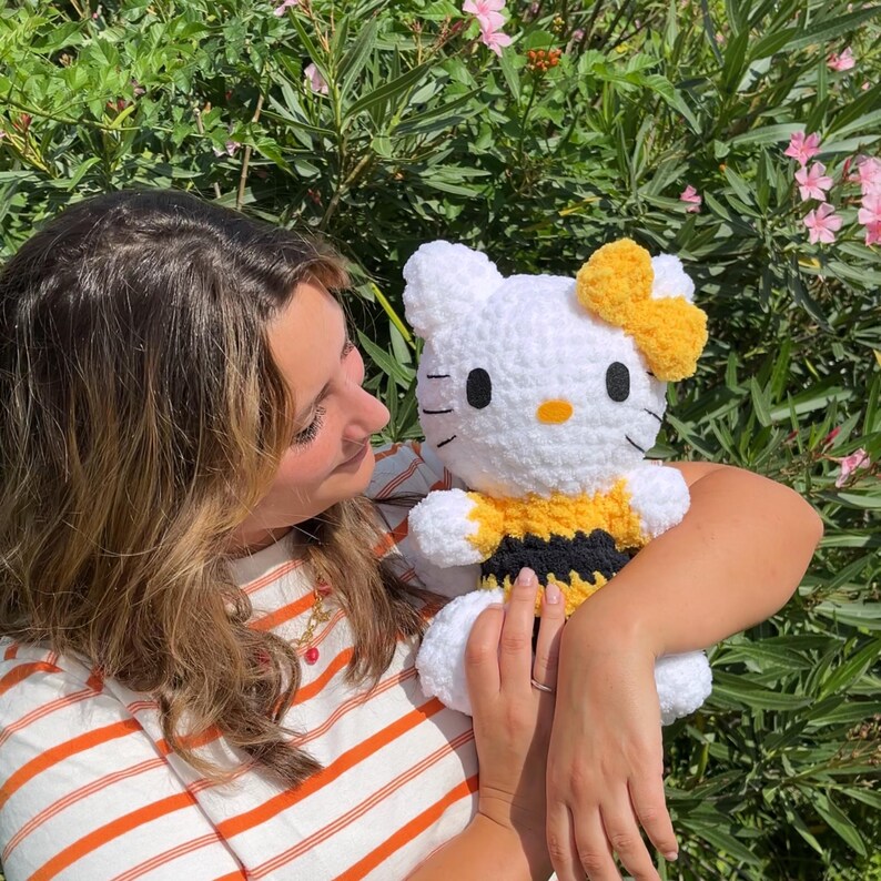 Puede incluir: Un juguete de peluche Hello Kitty blanco tejido a crochet con un disfraz de abeja amarillo y negro. El juguete est&aacute; siendo sostenido por una persona.