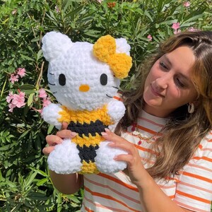 Puede incluir: Un juguete de peluche Hello Kitty blanco tejido a crochet con un disfraz de abeja amarillo y negro. El juguete tiene un lazo amarillo en la cabeza y est&aacute; siendo sostenido por una persona.