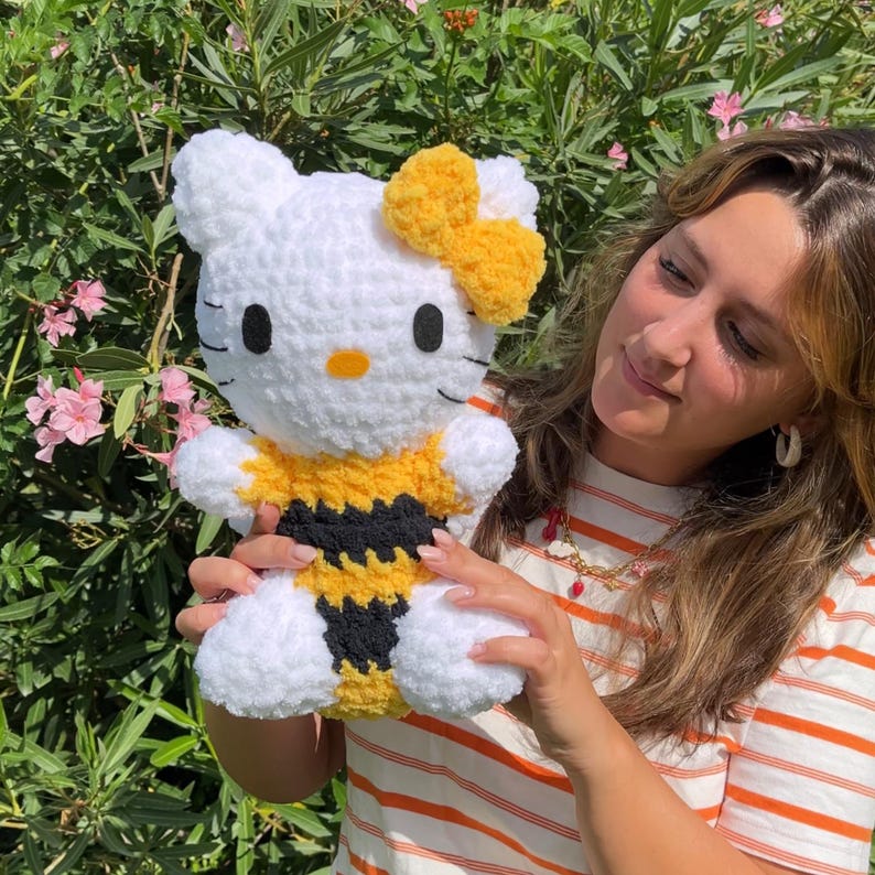 Puede incluir: Un juguete de peluche Hello Kitty blanco tejido a crochet con un disfraz de abeja amarillo y negro. El juguete tiene un lazo amarillo en la cabeza y est&aacute; siendo sostenido por una persona.