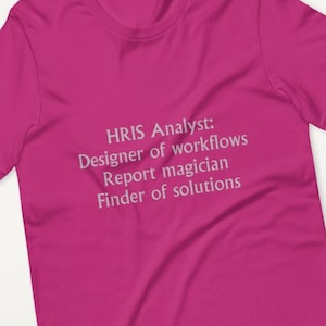 Może przedstawiać: Magenta koszulka z napisem "HRIS Analyst: Designer of workflows Report magician Finder of solutions" w jasnoszarej czcionce. Koszulka jest prezentowana na białej powierzchni z rozrzuconym konfetti i zielenią.