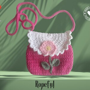 May include: A handmade pink crochet purse with a white scalloped flap and a pink flower applique. The purse has a long pink strap and a green stem and leaf design. The word "Ropeful!" is written at the bottom.