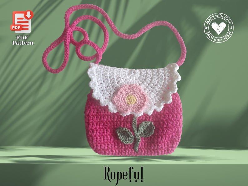 May include: A handmade pink crochet purse with a white scalloped flap and a pink flower applique. The purse has a long pink strap and a green stem and leaf design. The word "Ropeful!" is written at the bottom.