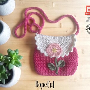 May include: A handmade pink crochet purse with a white scalloped top and a pink flower applique. The purse has a long pink strap and the word "Ropeful!" is written at the bottom. The image also includes a PDF pattern icon.