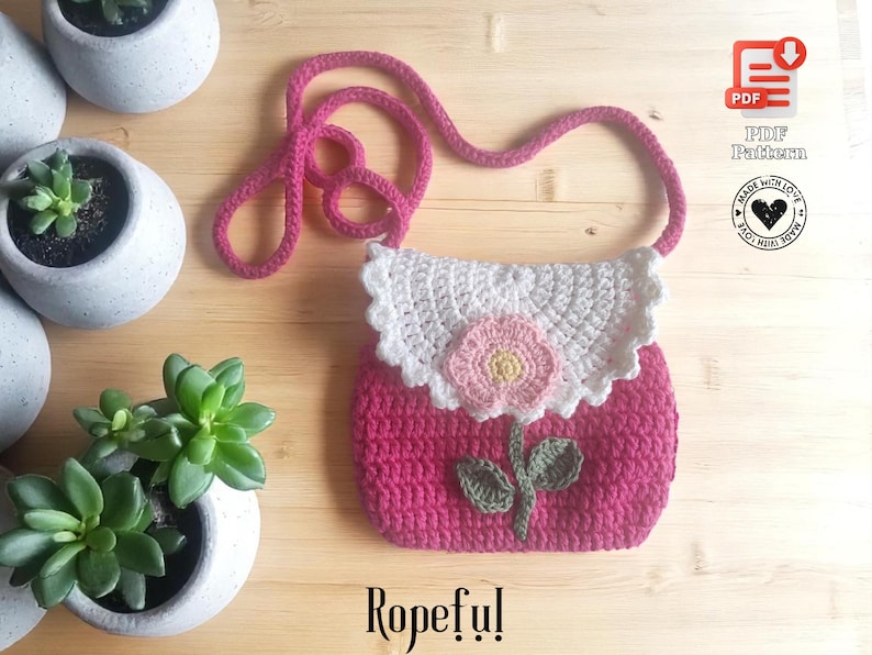 May include: A handmade pink crochet purse with a white scalloped top and a pink flower applique. The purse has a long pink strap and the word "Ropeful!" is written at the bottom. The image also includes a PDF pattern icon.