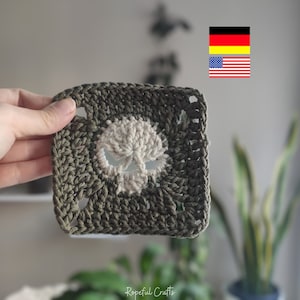 May include: A green crochet square with a white skull design in the center. The square is made with a granny stitch pattern.
