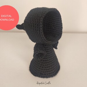 May include: Black crocheted amigurumi doll with a hooded head and a long, flowing robe. The doll is standing on a white surface.