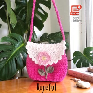 May include: A handmade pink crochet bag with a white scalloped flap and a pink flower applique. The bag has a long pink strap and a green leaf design. The word "Ropeful!" is written at the bottom.