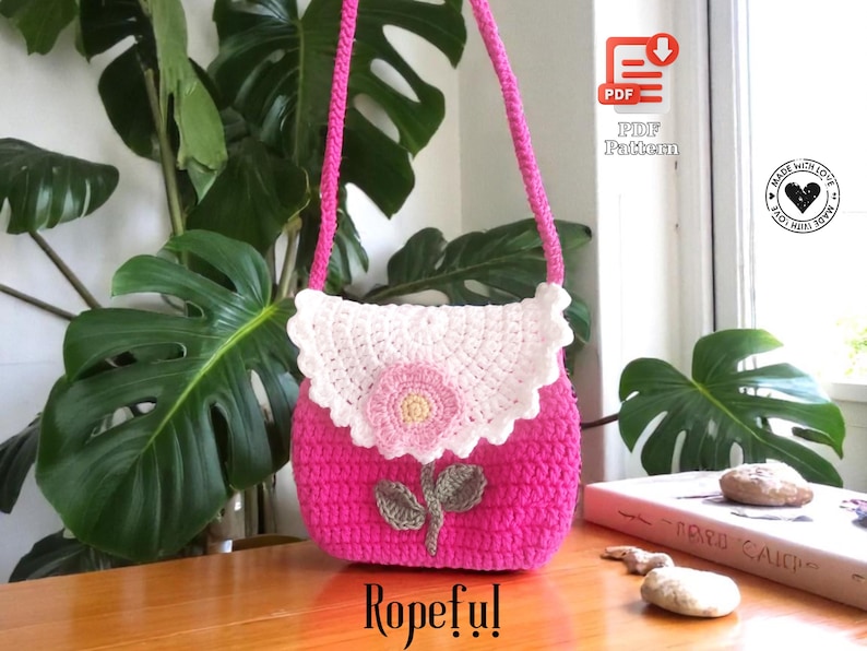 May include: A handmade pink crochet bag with a white scalloped flap and a pink flower applique. The bag has a long pink strap and a green leaf design. The word "Ropeful!" is written at the bottom.