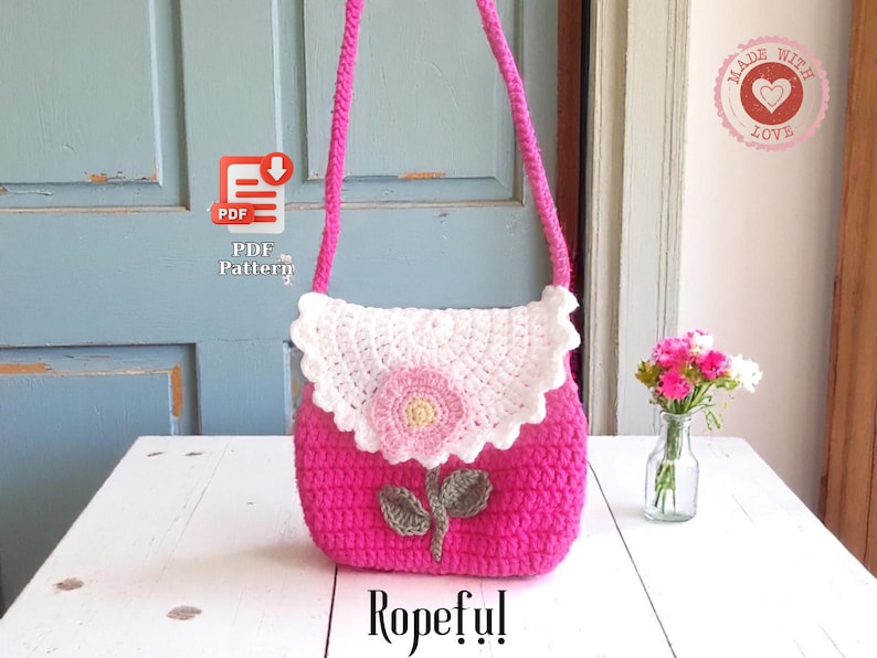 May include: A handmade pink crochet purse with a white scalloped flap and a pink flower applique. The purse has a long pink strap and a green leaf design. The word "Ropeful!" is written at the bottom.