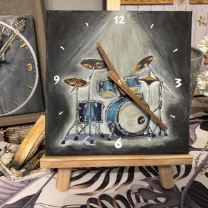 May include: A square wall clock featuring a painted illustration of a blue drum set on a dark gray background. The clock face displays white painted numbers at the twelve, three, six, and nine o'clock positions, with two wooden clock hands.
