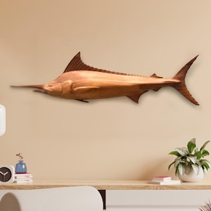 Op de afbeelding: Een houten beeldhouwwerk van een marlijn, gemonteerd aan een muur. De marlijn is bruin en heeft een lange, puntige bek.