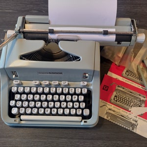 May include: A light blue Hermes 2000 typewriter with a white sheet of paper in the carriage. The typewriter is on a dark wood surface with two red instruction manuals.