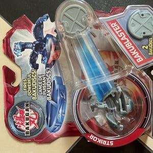 Peut inclure: Un ensemble de jouets Bakugan Striker dans un emballage en plastique transparent. L'emballage présente une palette de couleurs rouge et bleu avec le texte "BAKUBLASTER" et "STRIKOR". Le jouet est une figurine Bakugan grise et bleue avec un projectile bleu.