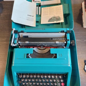May include: A teal Olivetti Studio 45 typewriter, open in its matching case. The keyboard has black keys with white lettering, and the carriage is silver. The case contains manuals, Tipp-Ex, and other accessories. The typewriter is on a dark wooden surface.