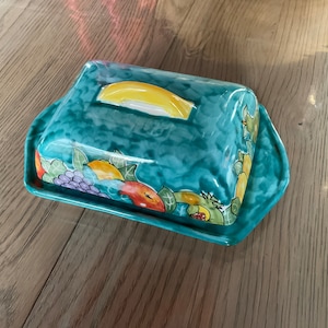 Peut inclure: Un beurrier en céramique bleu-vert avec un couvercle orné d'un motif de fruits colorés. Le couvercle a une poignée jaune.