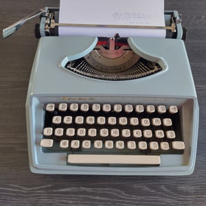 May include: A light blue typewriter with a white paper insert. The typewriter has a standard QWERTY keyboard with white keys and black lettering. The text on the paper reads: "Now I still wish we could go to the beach. I mean are you writing the script or just answering my questions.  I'm sure it's just a stalling tactic."