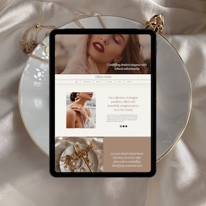 May include: A tablet displaying a website for "Hillary Robin" jewelry, showcasing designer pieces. The tablet rests on a white plate with a gold rim, surrounded by gold necklaces and a ring. The website features images of jewelry and text: "Combining timeless elegance with refined craftsmanship."