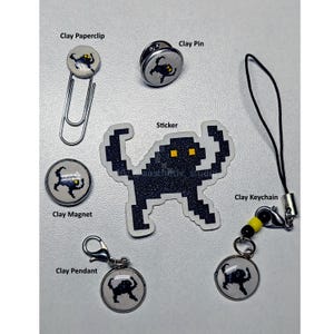May include: A collection of handmade clay accessories featuring a pixelated black creature design. Items include a paperclip, pin, magnet, sticker, keychain, and pendant. The accessories are on a light gray surface.