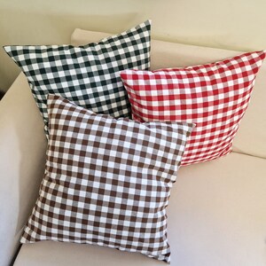 May include: Three square throw pillows in a variety of colors, arranged on a beige sofa. The pillows feature a classic gingham pattern in green and white, red and white, and brown and white. The pillows are soft and inviting.