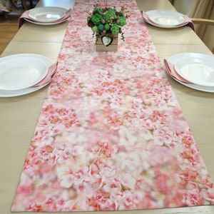 May include: A pink floral table runner with a blurred background. The runner features a delicate floral pattern with pink and white flowers. The runner is placed on a wooden table with two white plates and pink napkins on each side.