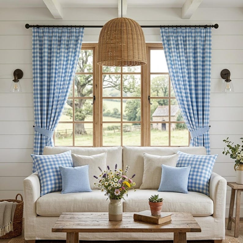 May include: A cozy living room scene featuring a cream-colored sofa adorned with blue and white gingham pillows. Blue gingham curtains frame a large window overlooking a scenic outdoor view. A woven pendant light hangs above a wooden coffee table.