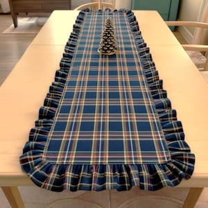 Pode incluir: Um caminho de mesa retangular com um padr&atilde;o xadrez em azul escuro, verde e vermelho, com uma borda de babados ao longo de todo o contorno. Um pequeno ornamento decorativo em forma de pinheiro c&oacute;nico prateado est&aacute; posicionado no centro.