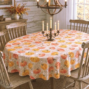 May include: An oval tablecloth featuring a repeating pattern of orange and yellow pumpkins on a light beige background with subtle script text. The fabric drapes over a dining table, which is set with a dark metal candelabra holding five white candles.