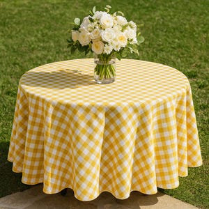 May include: A round table covered with a yellow and white checkered tablecloth. A glass vase with white roses and greenery sits in the center. The table is outdoors on a green lawn.