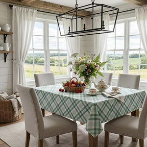 May include: A square dining table covered with a green and white plaid tablecloth, featuring a wicker basket filled with assorted fruits, a floral teapot, two teacups, and a glass vase containing a bouquet of wildflowers and greenery.