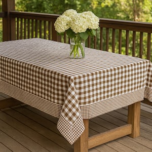 May include: A rectangular tablecloth featuring a brown and white gingham check pattern with a wider checkered border along the edges. A clear glass vase containing a large bouquet of white hydrangea flowers sits on the center of the table.