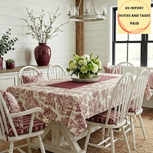 Peut inclure: Une salle à manger avec une table et des chaises blanches. La table est recouverte d'une nappe crème avec un motif de toile rouge. Des coussins rayés rouges et blancs ornent les chaises. Un vase bordeaux et une composition florale sont sur la table.