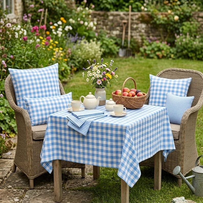 May include: A charming outdoor setting with wicker chairs, a table, and a garden backdrop. The table is covered with a blue and white checkered tablecloth and set with a teapot, cups, and a basket of fruit. Blue checkered pillows adorn the chairs.