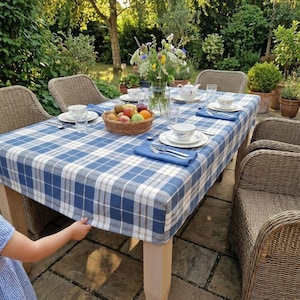 May include: A rectangular dining table covered with a blue and white plaid tablecloth, surrounded by woven wicker chairs. The table is set with white ceramic plates, blue napkins, clear glasses, a fruit basket, and a glass vase of wildflowers.