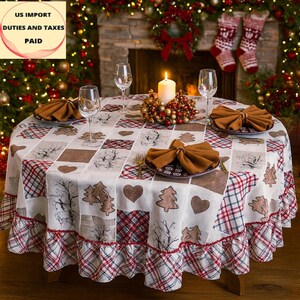 Op de afbeelding: Een ronde tafelkleed met een patchwork ontwerp met bomen, harten en ruitpatronen in rood, bruin en wit. De tafel is gedekt met borden, servetten en wijnglazen, met een brandende kaars en feestelijke decoraties op de achtergrond.