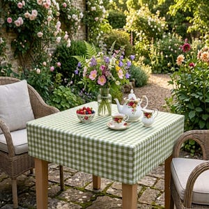 May include: A square table covered with a green and white gingham tablecloth, featuring a glass vase of colorful wildflowers, a white porcelain teapot with floral patterns, a matching teacup on a saucer, and a small bowl filled with fresh berries.