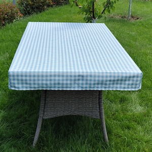 Puede incluir: Un mantel de mesa a cuadros azules y blancos sobre una mesa de mimbre gris. El mantel está hecho de una tela ligera y tiene un diseño sencillo.