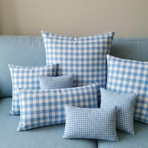 Puede incluir: Un conjunto de cojines a cuadros vichy azules y blancos de varios tamaños, colocados sobre un sofá azul claro. Los cojines tienen una estética clásica y campestre, ideal para la decoración del hogar.