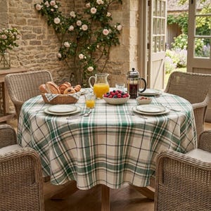 May include: A round table covered with a green, white, and tan plaid tablecloth, set with a wicker basket of bread, a glass pitcher of orange juice, a bowl of fresh berries, a glass French press, and white ceramic plates with silverware.