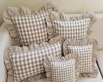 May include: A collection of five square and rectangular throw pillows featuring a beige and white gingham check pattern. Each pillow is finished with a gathered fabric ruffle border around the entire perimeter, creating a consistent look.